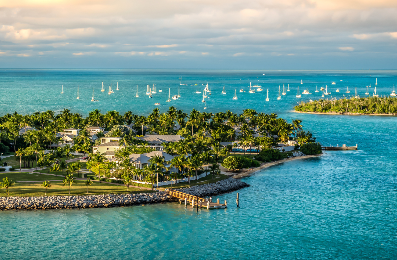 An aerial view over Key West, Florida with bright blue waters surrounding lush green islands and white sailboats in the distance.