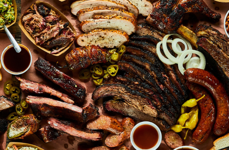 A table full of smoked ribs, sausage, smoked chicken, pulled pork, and brisket, with sliced onions and peppers