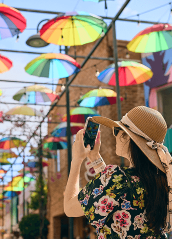 A lady in a sun hat taking a photo with her phone of her surroundings: brightly colored umbrellas hanging over the street of Dominican Republic.