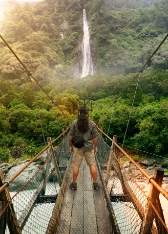 A person standing on a bridge while staring at a waterfall in the distance among the jungle of trees.