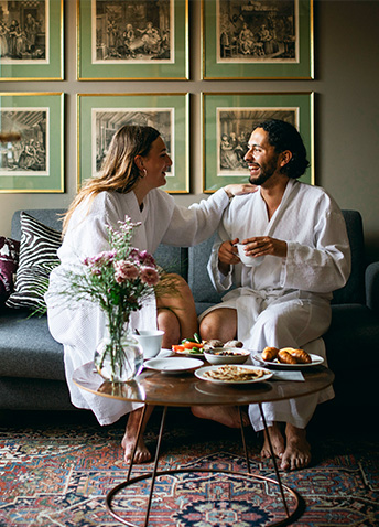 A young woman and young man wearing white fluffy robes enjoying their hotel breakfast on the coffee table in the comfort of their room while sitting on the couch