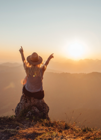 A woman in a hat cheering towards the sunset.