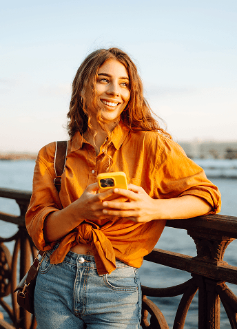 A smiling young woman holding a yellow smartphone while leaning against a railing near a body of water at sunset.