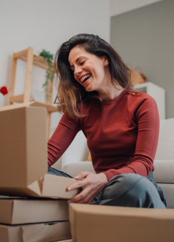A woman sitting on the floor smiles as she opens cardboard delivery boxes in her living room.