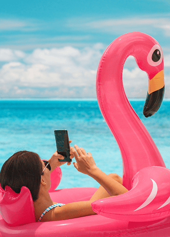 A woman using her smartphone while relaxing on an inflatable pink flamingo float in tropical turquoise ocean waters.