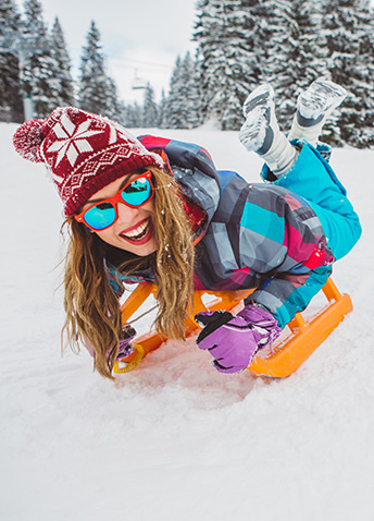 A smiling woman in colorful winter gear and a red patterned beanie lies on an orange sled in the snow.