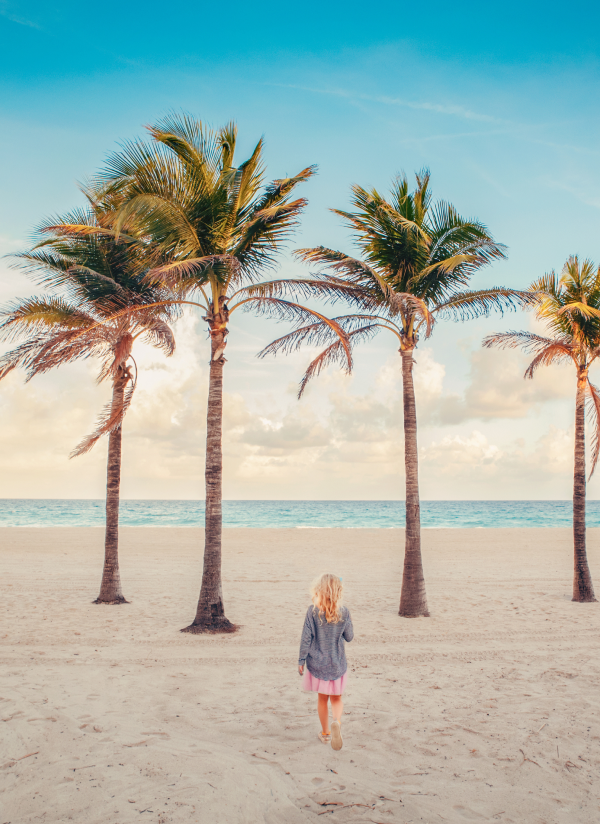A woman walking on a sandy beach dotted by tall palm trees with the ocean visible in the background.