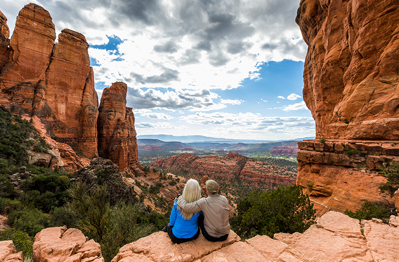A senior couple sits arm-in-arm on a red rock ledge, looking out over a vast, scenic canyon landscape in Sedona, Arizona under a dramatic cloudy blue sky.