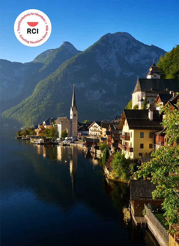Scenic view of a European alpine village on a calm lake with tall church steeples set against steep green mountains.