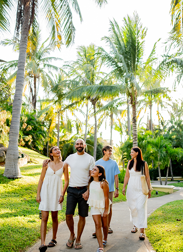 A happy family of five walks together smiling along a paved path lined with lush palm trees at a tropical resort destination.