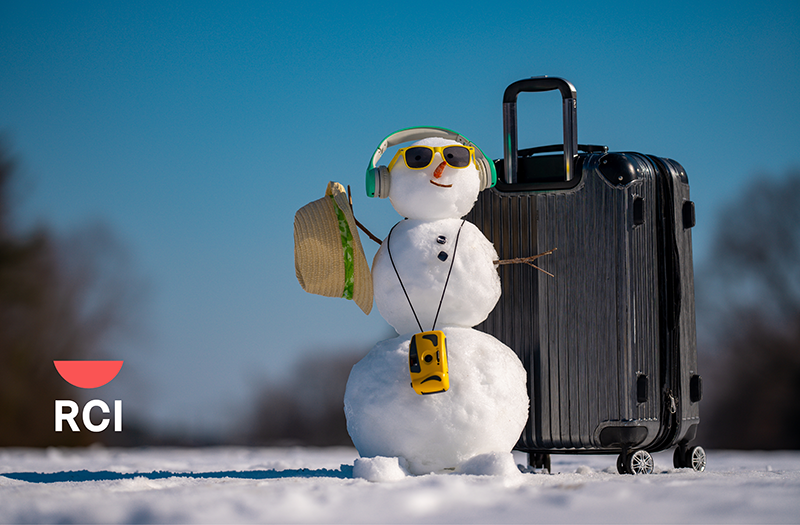 A small snowman wearing sunglasses, headphones, and a camera, standing next to a large black suitcase in the snow.