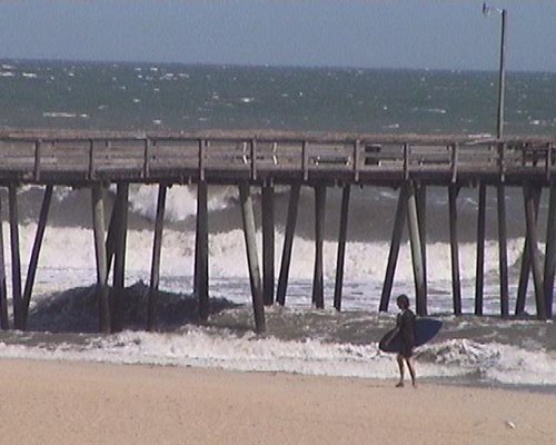 Surfer walking on a beach with a wooden pier and ocean-view.