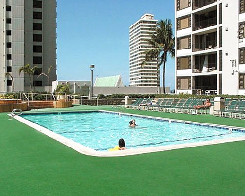 An outdoor turf edged swimming pool with chaise lounge chairs alongside multi story condos balconies.