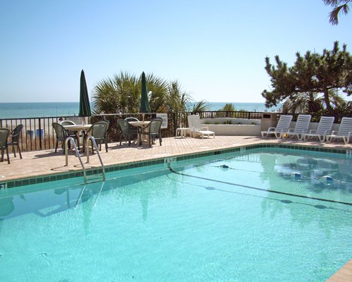 Outdoor pool with chaise lounge chairs and patio tables alongside ocean.