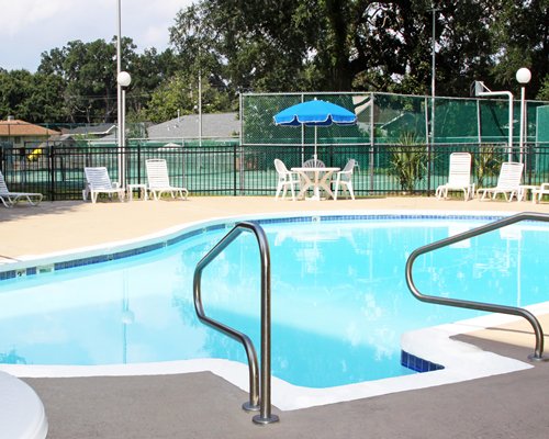 Outdoor swimming pool with chaise lounge chairs in front of a tennis court surrounded by wooded area.