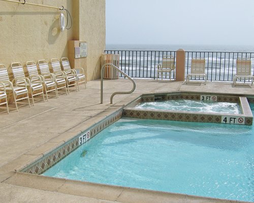 An outdoor swimming pool and hot tub with chaise lounge chairs along the shore.