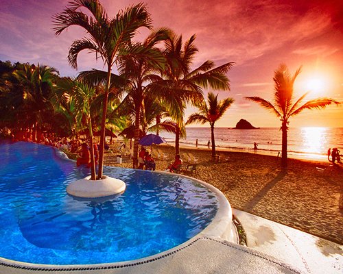 Dusk view of outdoor swimming pool with thatched sunshades and patio furniture alongside the beach.