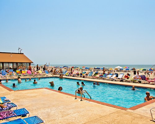 View of people at an outdoor swimming pool with chaise lounge chairs and sunshades alongside the ocean.