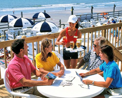 A steward serves the cocktail to the family alongside chaise lounge chairs sunshades facing the ocean.