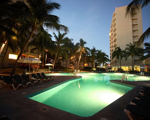 An outdoor swimming pool with chaise lounge chairs and palm trees with view of multiple units at dusk.