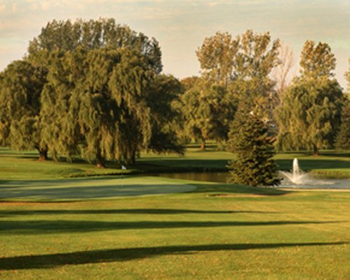 View of the golf course and pond with fountain surrounded by wooded area.