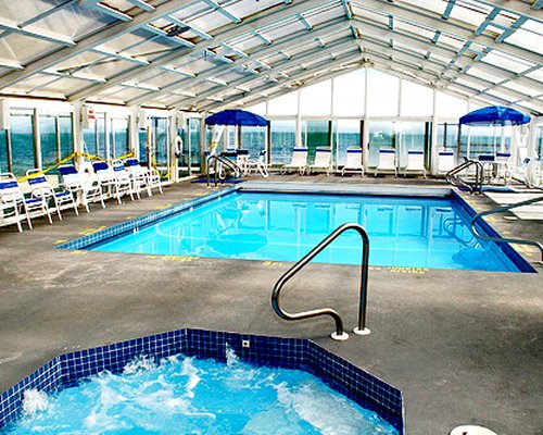 Indoor swimming pool with an indoor hot tub chaise lounge chairs sunshades and outside view.