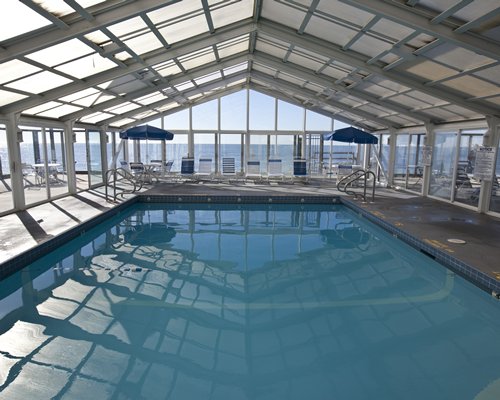 An indoor swimming pool with chaise lounge chairs patio with sunshade and an outside view.