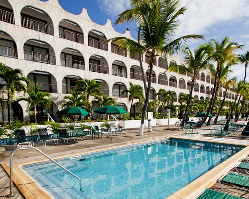 Outdoor swimming pool with chaise lounge chairs sunshades and palm trees alongside multiple balconies.