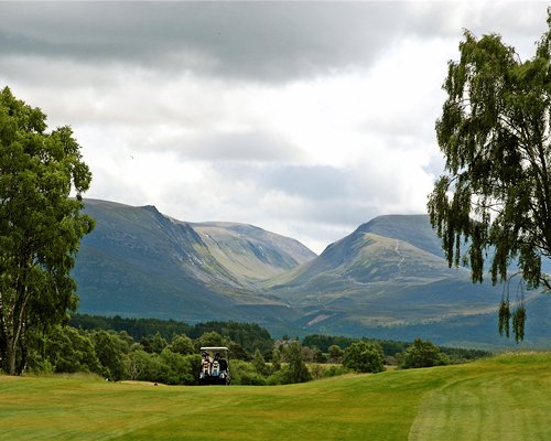 View of the golf course with a golf cart alongside wooded area and mountains.