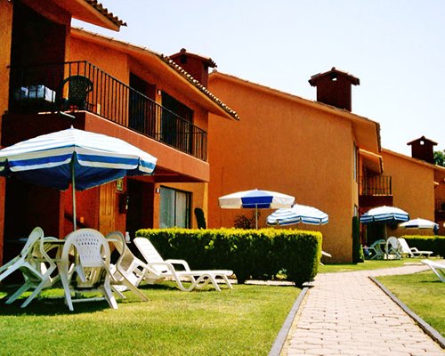 Scenic exterior view a unit with balcony alongside a pathway with patio chairs and sunshades.