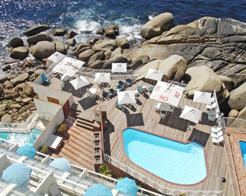 An aerial view of the swimming pool with shaded chaise lounge chairs alongside the ocean.