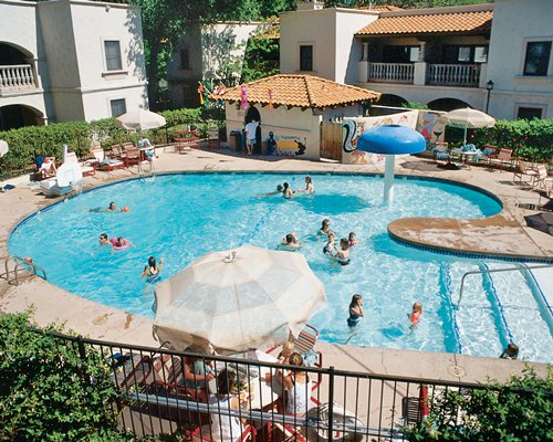 An outdoor swimming pool with a raining mushroom umbrella alongside multi story resort units.