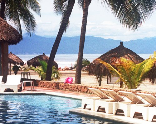 Outdoor swimming pool with chaise lounge chairs alongside the beach with palm trees and thatched sunshades.