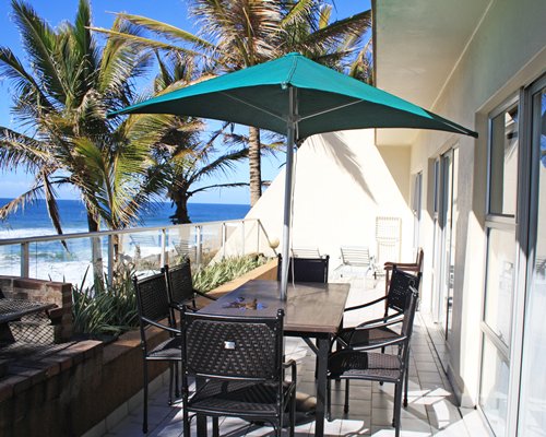 Balcony with sunshade and patio chairs alongside the ocean with palm trees.