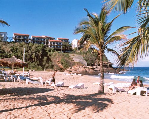 View of Resort Caravelas alongside the beach with chaise lounge chairs thatched sunshade and palm trees.