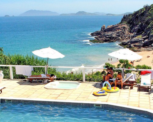 An outdoor swimming pool with hot tub chaise lounge chairs and thatched sunshades alongside the ocean.