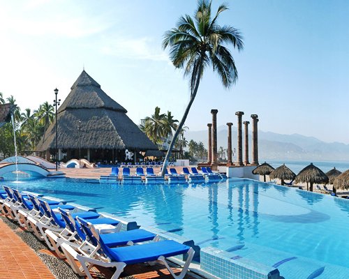Outdoor swimming pool with chaise lounge chairs thatched sunshades and palm trees alongside the beach.