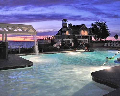 A large outdoor swimming pool with chaise lounge chairs alongside resort units at dusk.