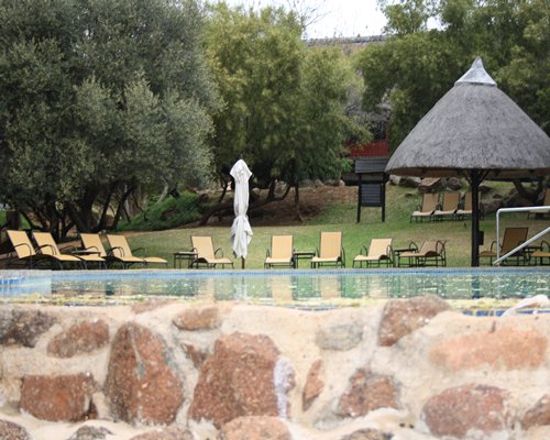 A scenic outdoor swimming pool with chaise lounge chairs and thatched sunshades surrounded by wooded area.