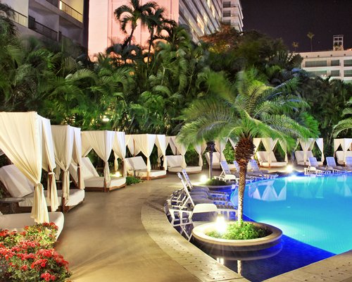 An outdoor swimming pool with chaise lounge chairs beach beds and palm trees at dusk.