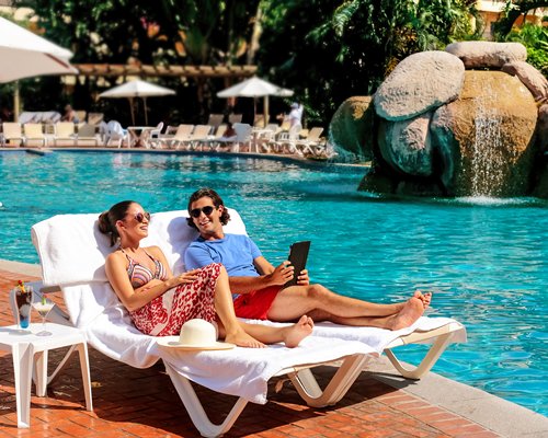 A couple relaxing on chaise lounge chairs alongside an outdoor swimming pool with sunshades.