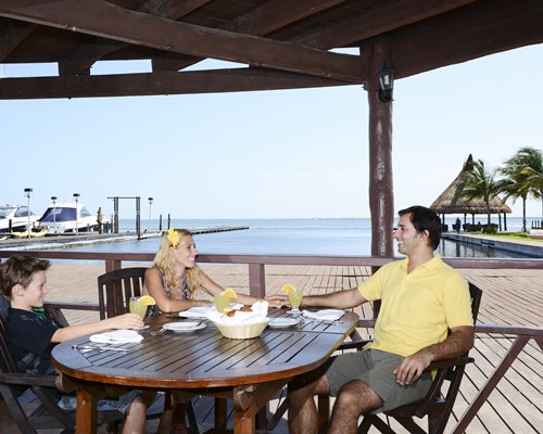 Family at outdoor restaurant alongside beach with boats and thatched sunshades.