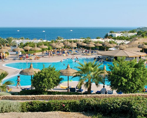 Outdoor swimming pool with chaise lounge chairs and thatched sunshades alongside the sea.