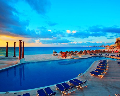An outdoor swimming pool with chaise lounge chairs and thatched sunshades alongside the ocean.