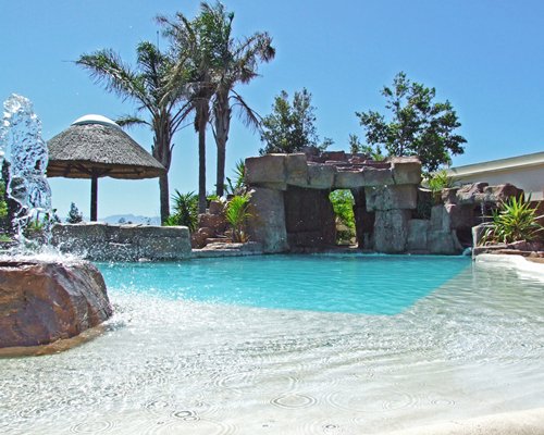 An outdoor swimming pool with water fountain alongside thatched sunshade.
