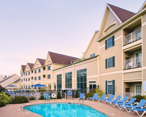 SCENIC EXTERIOR VIEW OF WYNDHAM BENTLEY BROOK WITH OUTDOOR SWIMMING POOL CHAISE LOUNGE CHAIRS AND SUNSHADES.