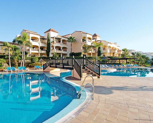 An outdoor swimming pool with thatched sunshades and chaise lounge chairs alongside a resort.