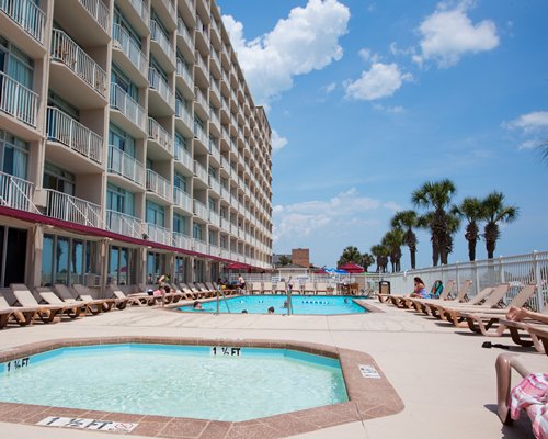 An outdoor swimming pool with a hot tub and chaise lounge chairs alongside multi story resort units.