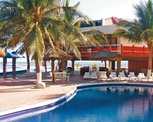 An outdoor swimming pool with chaise lounge chairs and thatched sunshades alongside the ocean.