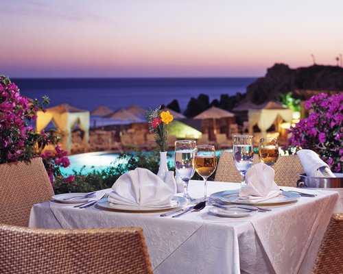 An outdoor dining area with beverages alongside the pool with thatched sunshades and the ocean.